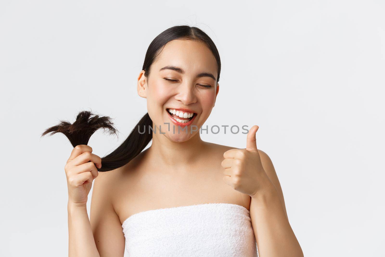 Beauty, hair loss products, shampoo and hair care concept. Close-up of satisfied, happy asian girl in bath towel showing thumbs-up and healthy hair ends, standing pleased white background.