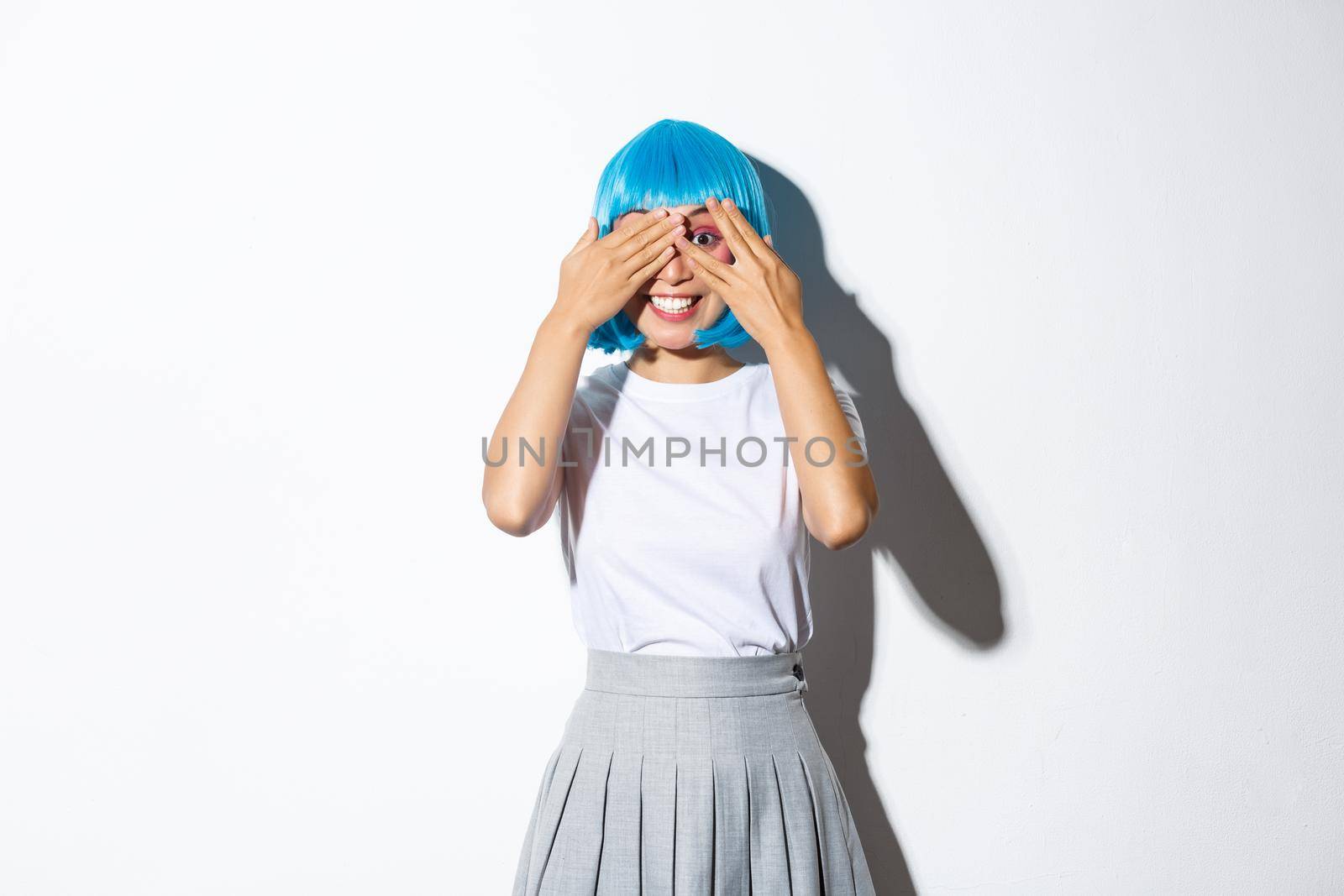 Portrait of dreamy cute asian girl in blue wig close eyes and peeking at camera with happy smile, standing over white background in halloween costume.