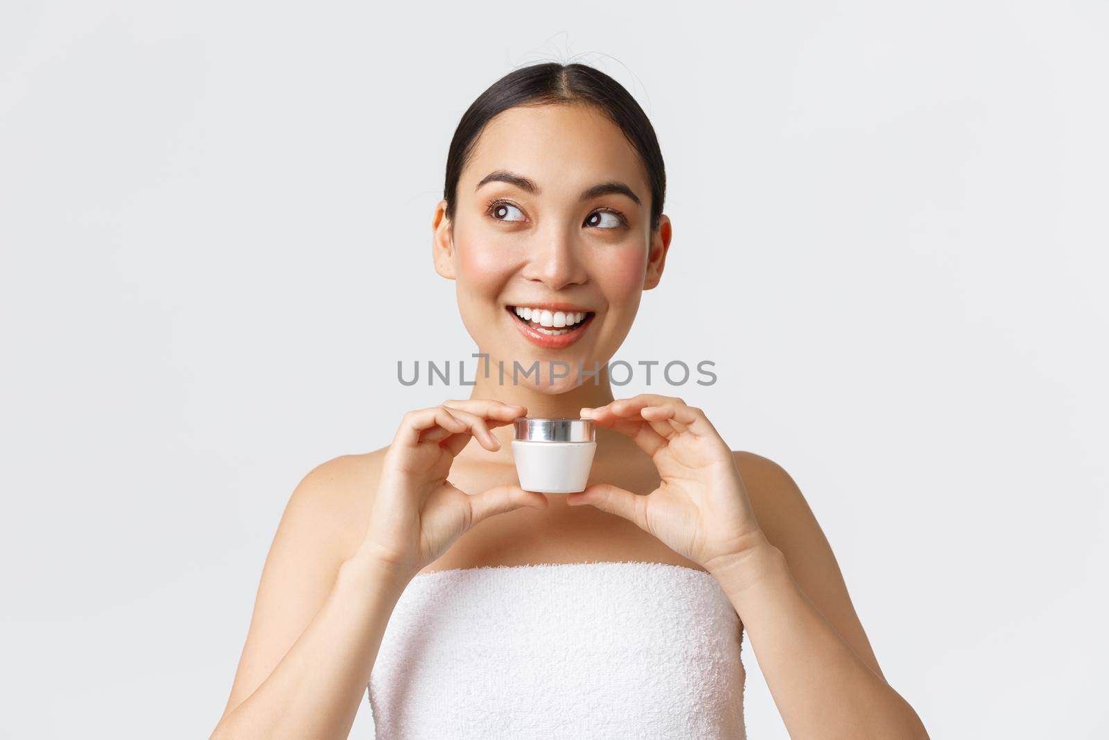Beauty, personal care, spa salon and skincare concept. Excited beautiful asian woman in bath towel showing face cream, looking upper left corner and smiling, recommend skin care product.