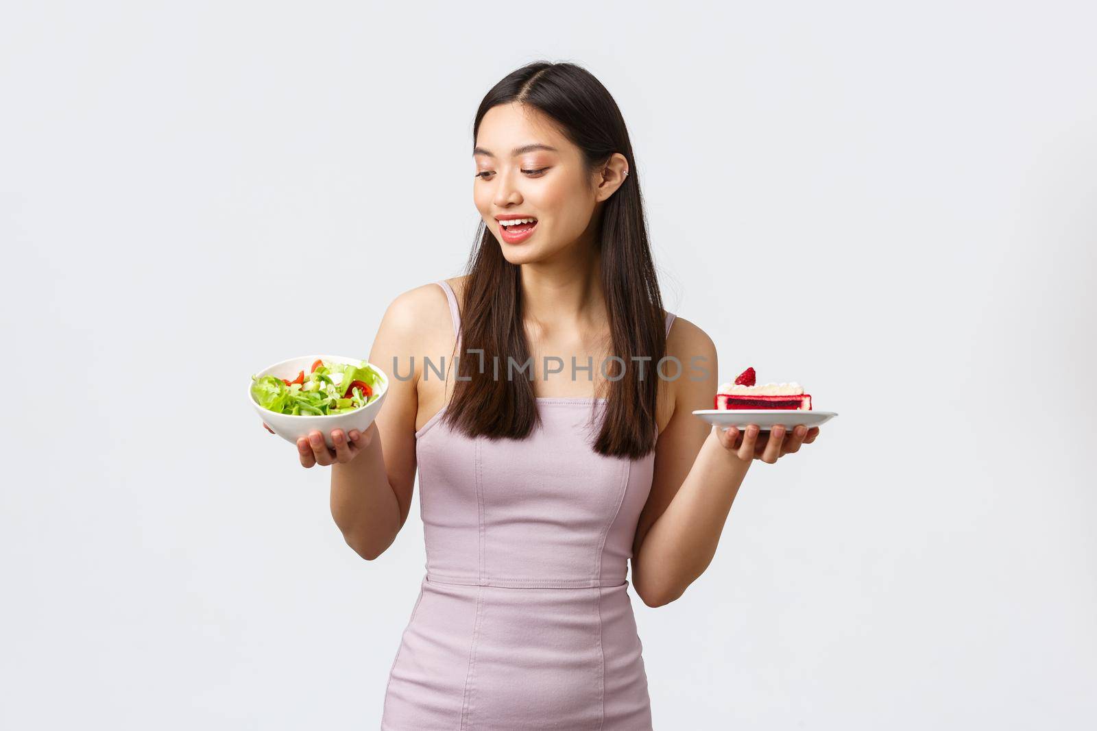 Healthy lifestyle, leisure and food concept. Beautiful slim asian girl in evening party dress, looking pleased at bowl with salad and holding piece of cake, standing white background.