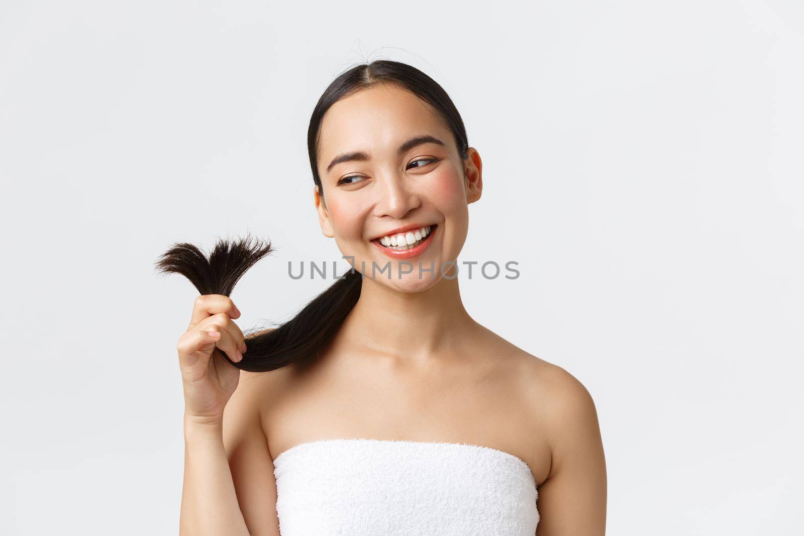 Beauty, hair loss products, shampoo and hair care concept. Gorgeous happy asian woman in bath towel showing hair ends and smiling satisfied, cured split ends and looking satisfied.