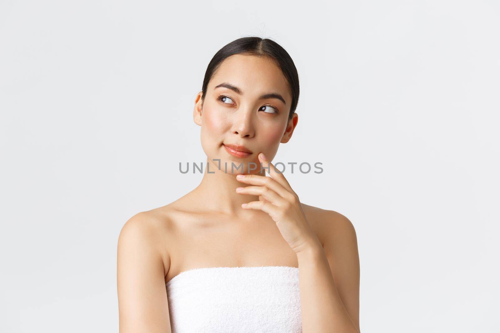 Beauty, cosmetology and spa salon concept. Thoughtful beautiful asian woman in towel looking left and thinking, pondering or making choice which procedure take in beauty clinic.