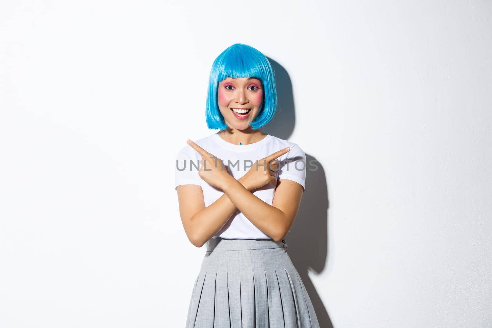 Happy beautiful asian girl turn attention to two promo banners, pointing fingers sideways and smiling amazed, standing in blue wig, dressed-up for party or halloween.