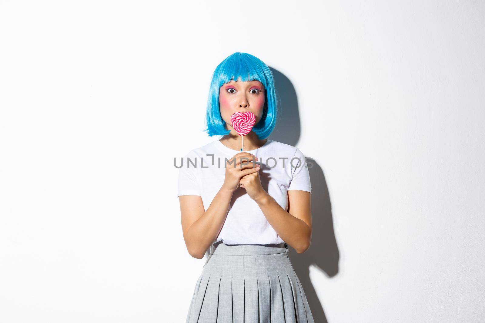 Image of coquettish asian girl in blue short wig, kissing heart-shaped candy, celebrating halloween, standing over white background.