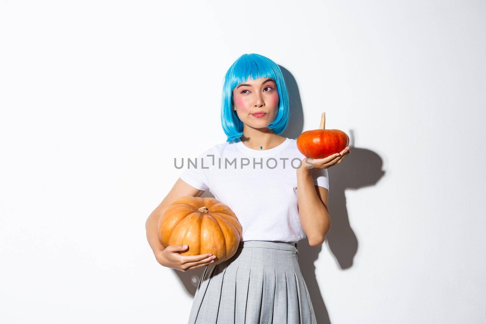 Portrait of thoughtful cute asian woman looking away while making choice, holding two different pumpkins, decorating halloween party, wearing blue wig.