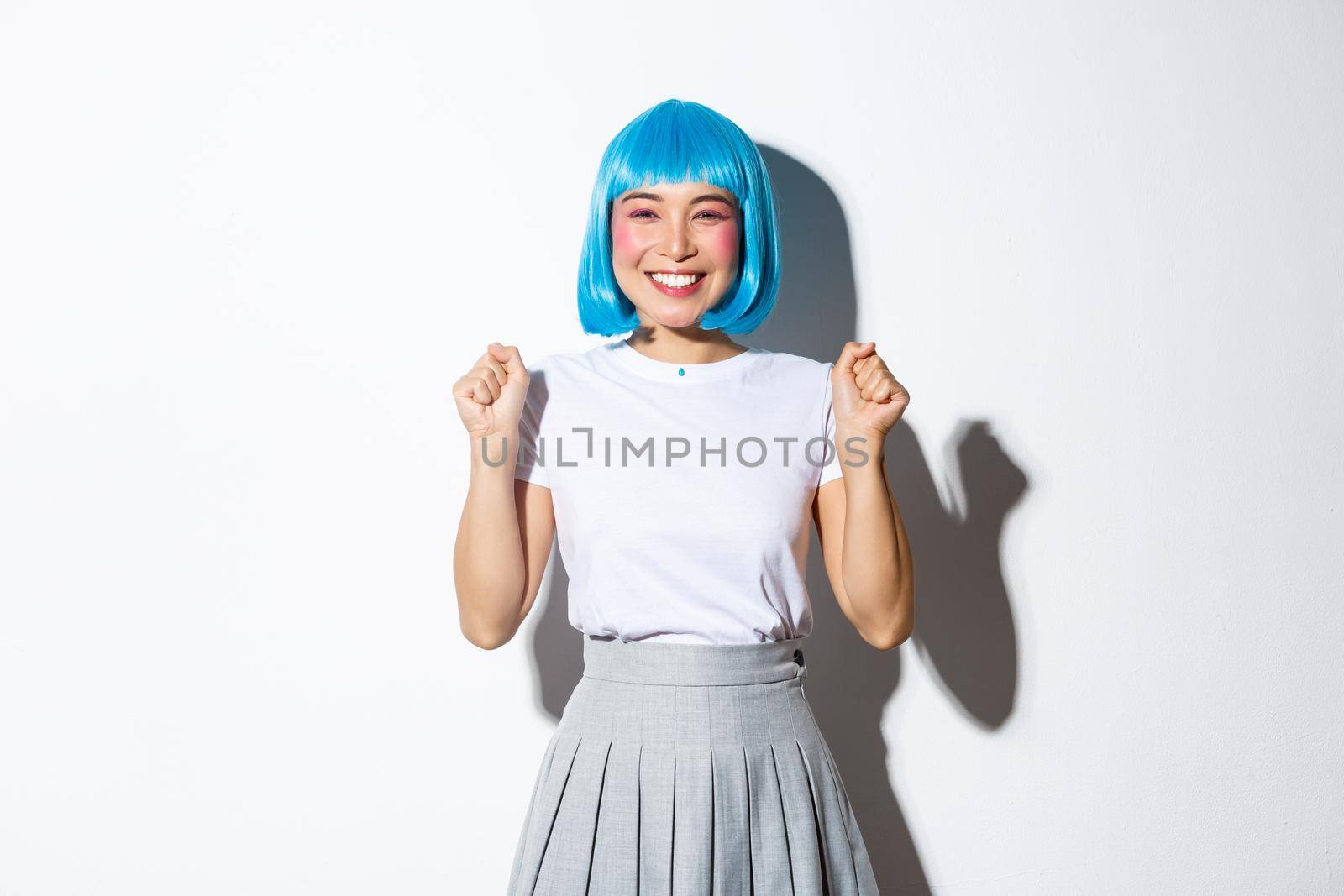 Portrait of cheerful kawaii asian girl in blue party wig celebrating victory, smiling happy and jumping from rejoice, triumphing over success, standing over white background.