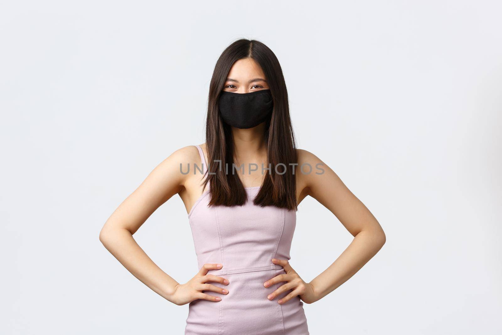 Covid-19, social distancing quarantine and leisure concept. Gorgeous and stylish asian woman in face mask, protect herself from coronavirus, attend party in elegant dress, white background.