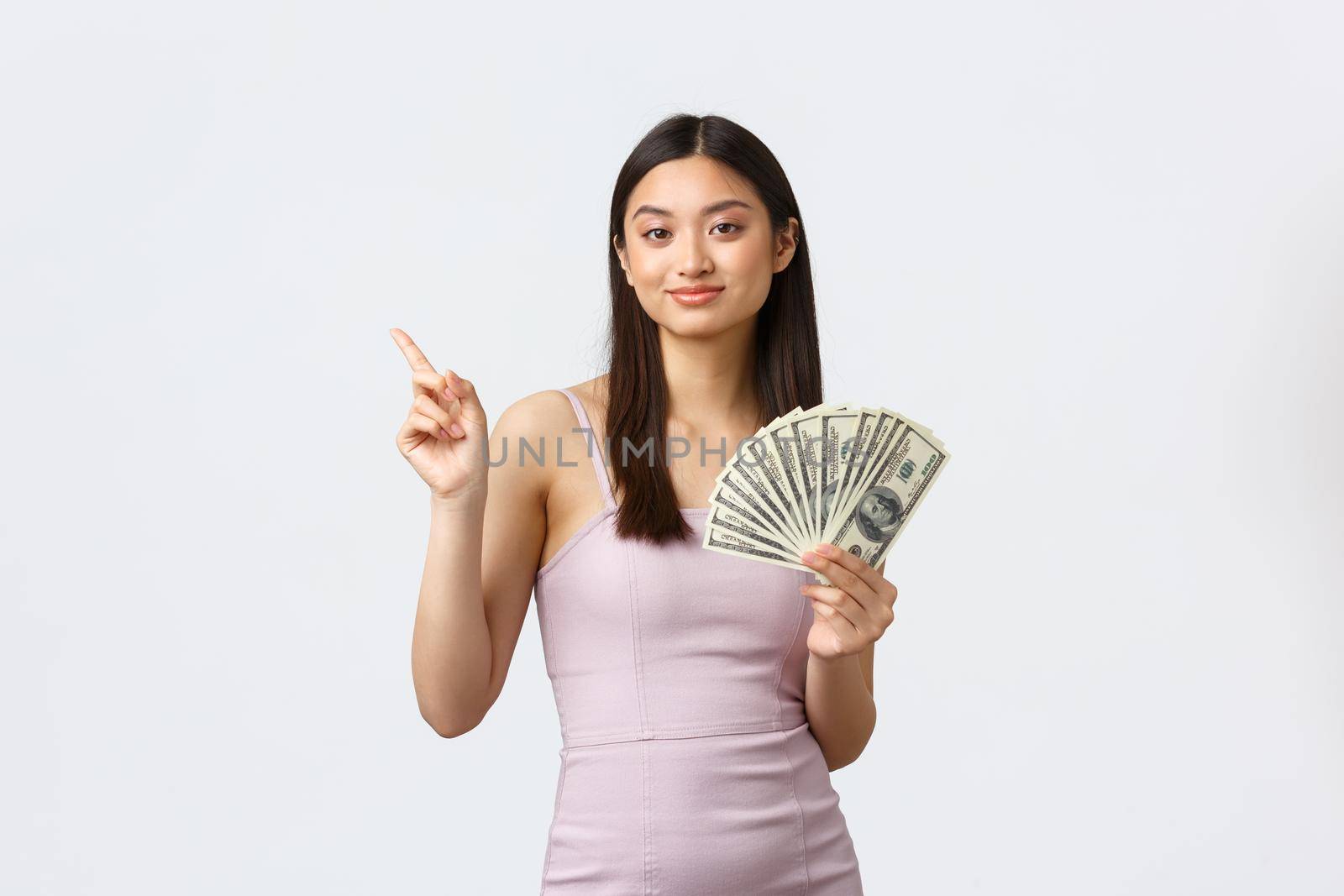 Shopping, holidays and people emotions concept. Cheerful smiling, happy korean woman in evening dress, showing money and pointing finger upper left at banner, white background.