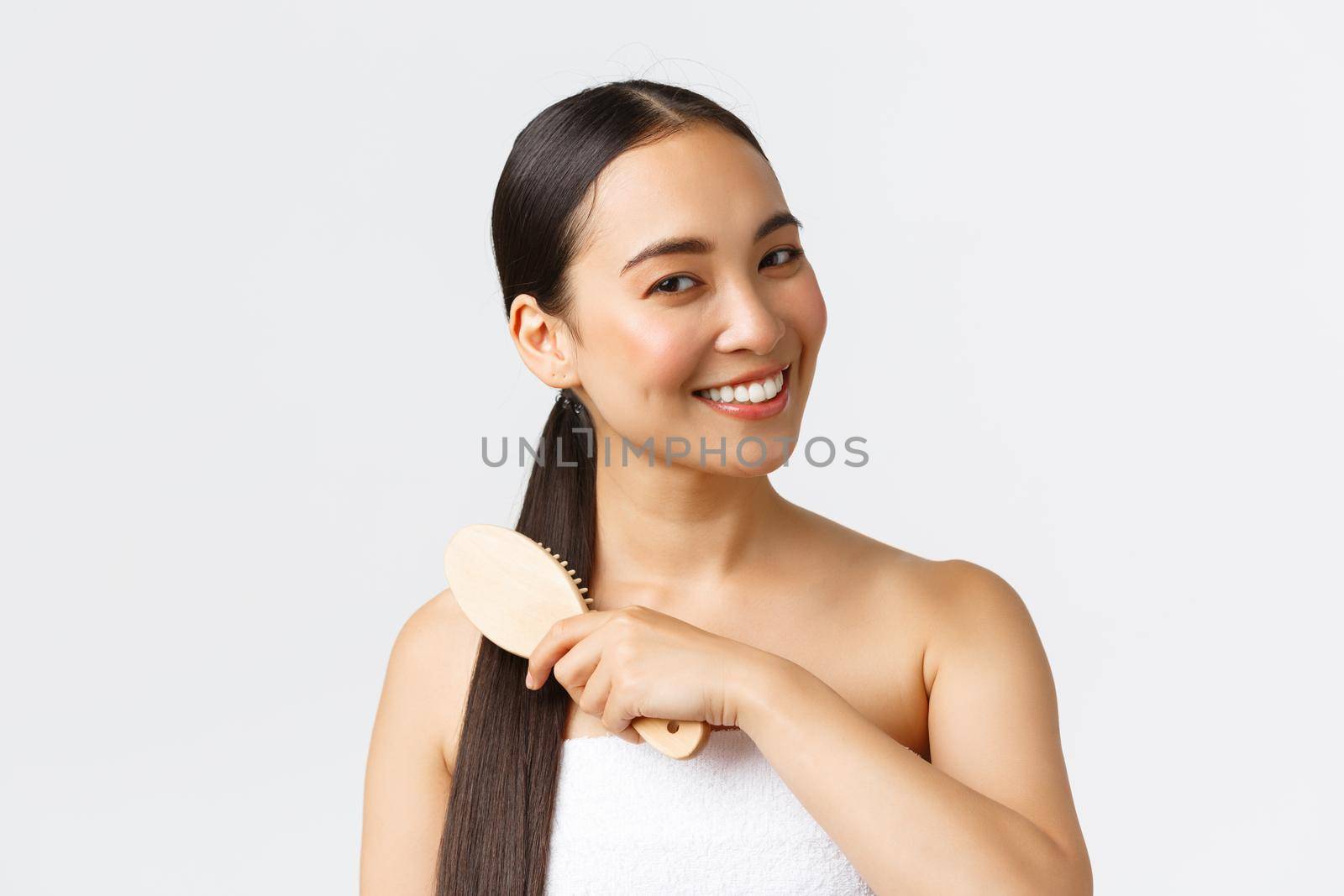 Beauty, hair loss products, shampoo and hair care concept. Close-up of beautiful asian female in bath towel brushing hair with brush and smiling, standing in bathroom over white background by Benzoix
