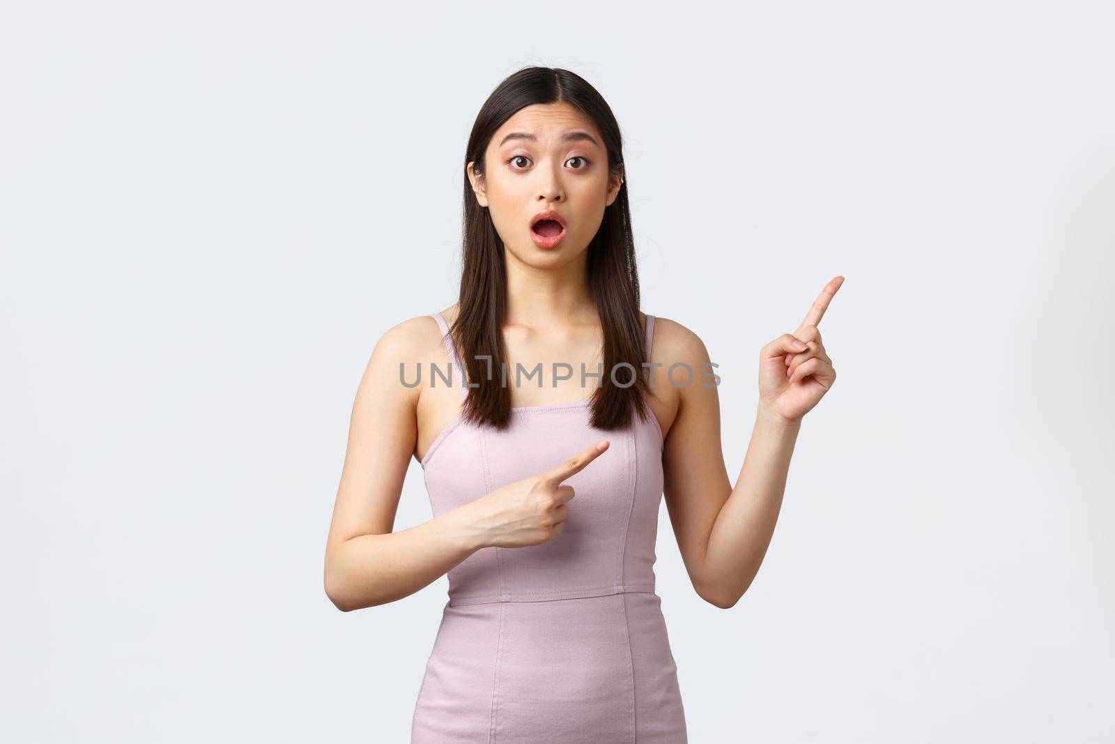 Luxury women, party and holidays concept. Excited and worried cute asian woman asking question or advice about event, wear evening dress and pointing fingers upper right corner.