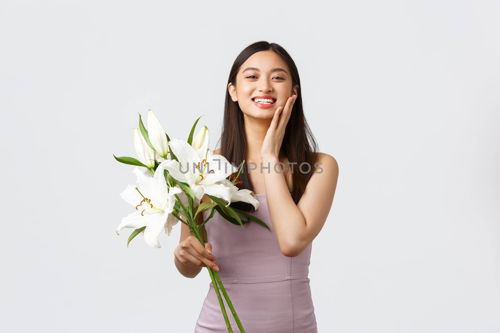 Holidays and events, celebration concept. Happy asian woman in evening dress, blushing and smiling surprised as receive bouquet of beautiful lilies, standing white background cheerful.