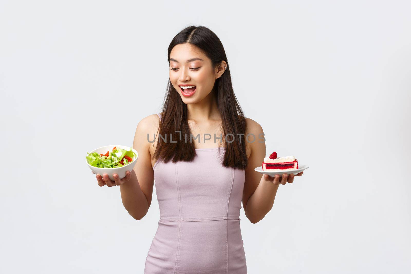 Healthy lifestyle, leisure and food concept. Cheerful beautiful asian girl in dress, looking excited with happy smile at salad instead of sweet cake, standing white background.