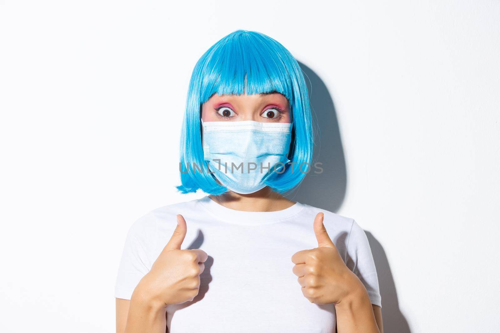 Concept of halloween celebration and coronavirus. Close-up of excited asian girl in medical mask and blue wig showing thumbs-up and approve something good by Benzoix