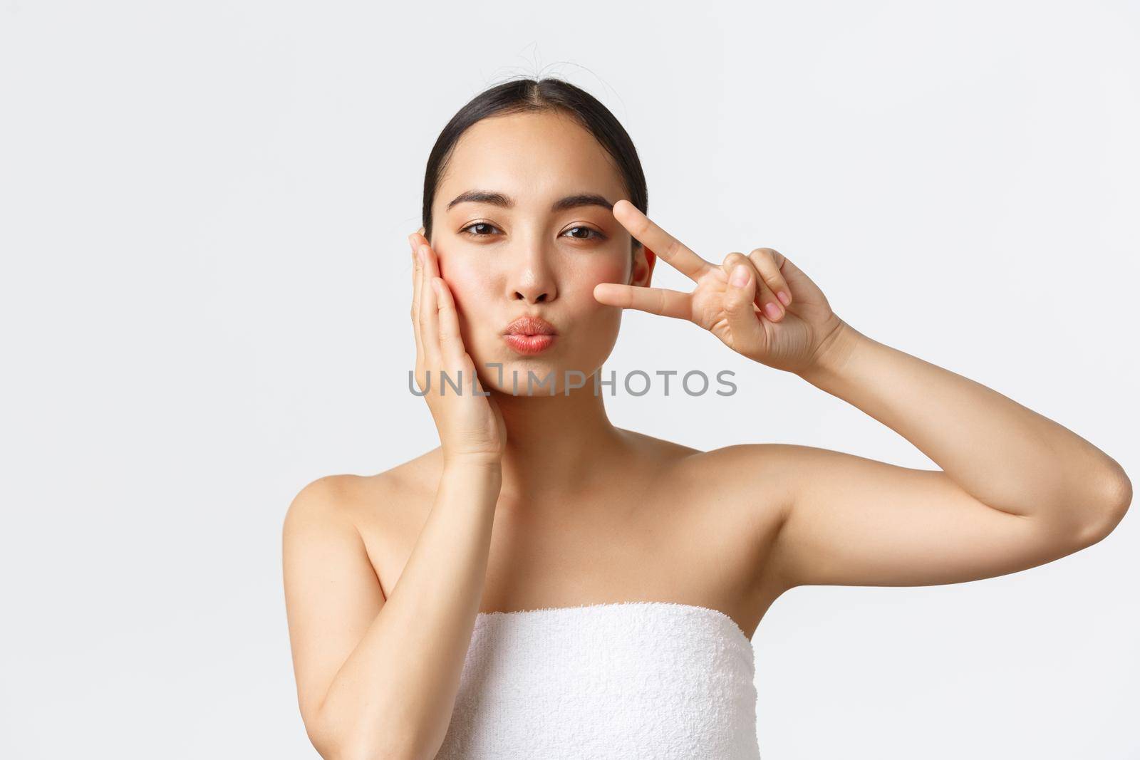 Beauty, personal care, spa salon and skincare concept. Close-up of sensual pretty asian girl in bath towel showing peace kawaii gesture and blowing air kiss, satisfied with skin condition by Benzoix