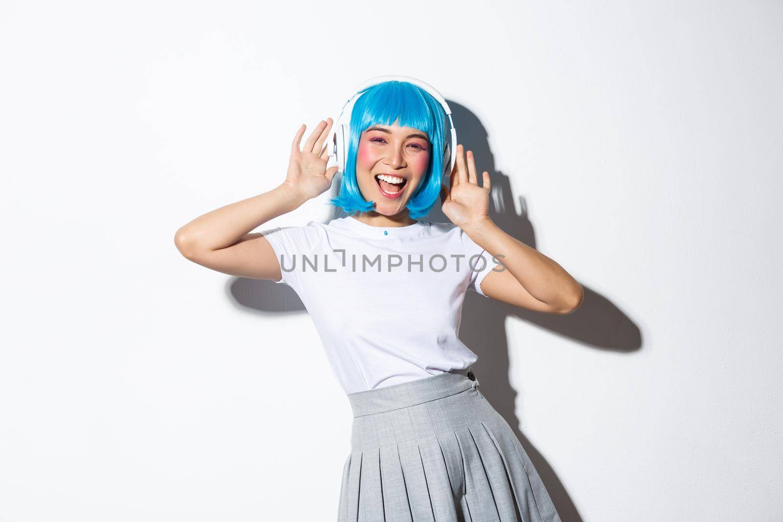 Cheerful asian girl in blue anime wig dancing and listening music in headphones, wearing halloween costume, standing over white background.