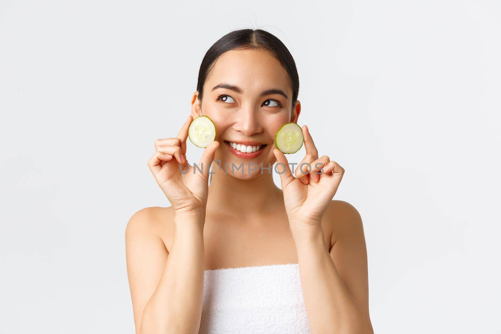 Beauty, personal care, spa salon and skincare concept. Beautiful young asian female in bath towel holding cucumbers and smiling, promo of facial or body treatment, moisturizing features of cream.