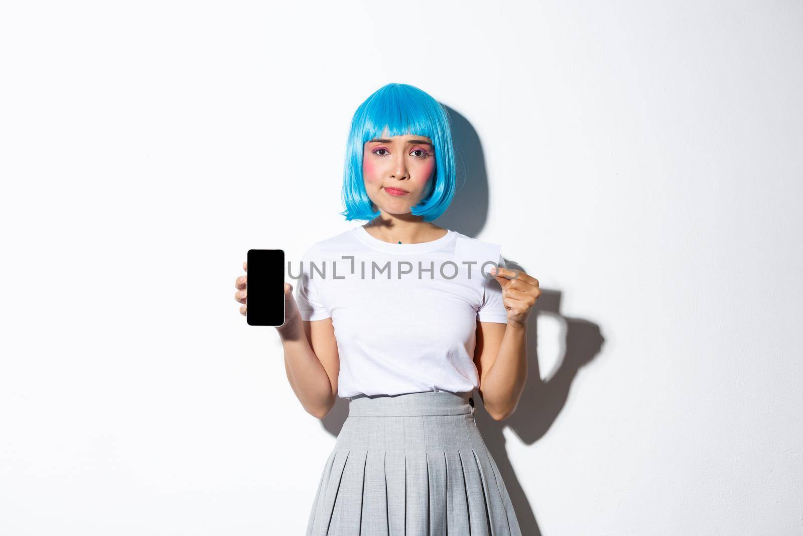 Portrait of skeptical asian girl in blue party wig, looking disappointed as showing credit card and smartphone screen, standing over white background.