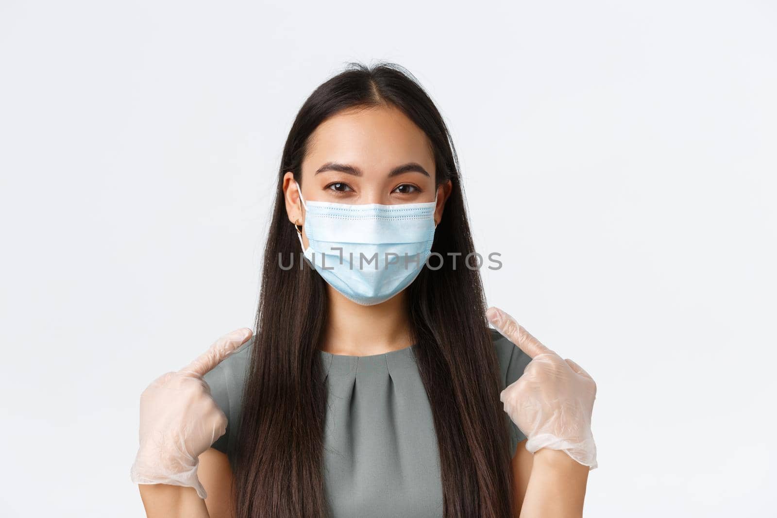 Small business owners, covid-19, preventing virus measures concept. Close-up of smiling asian woman in medical mask and gloves pointing at face, using protective equipment during pandemic.