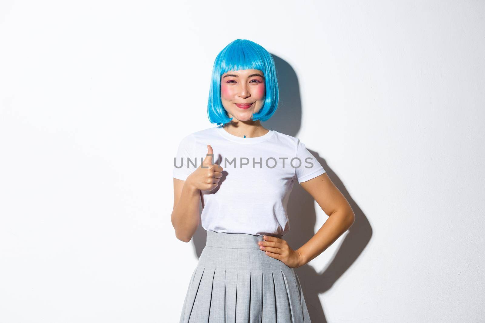 Portrait of cute asian girl in blue wig looking satisfied, smiling and showing thumbs-up in approval, dressed for halloween celebration, standing over white background.
