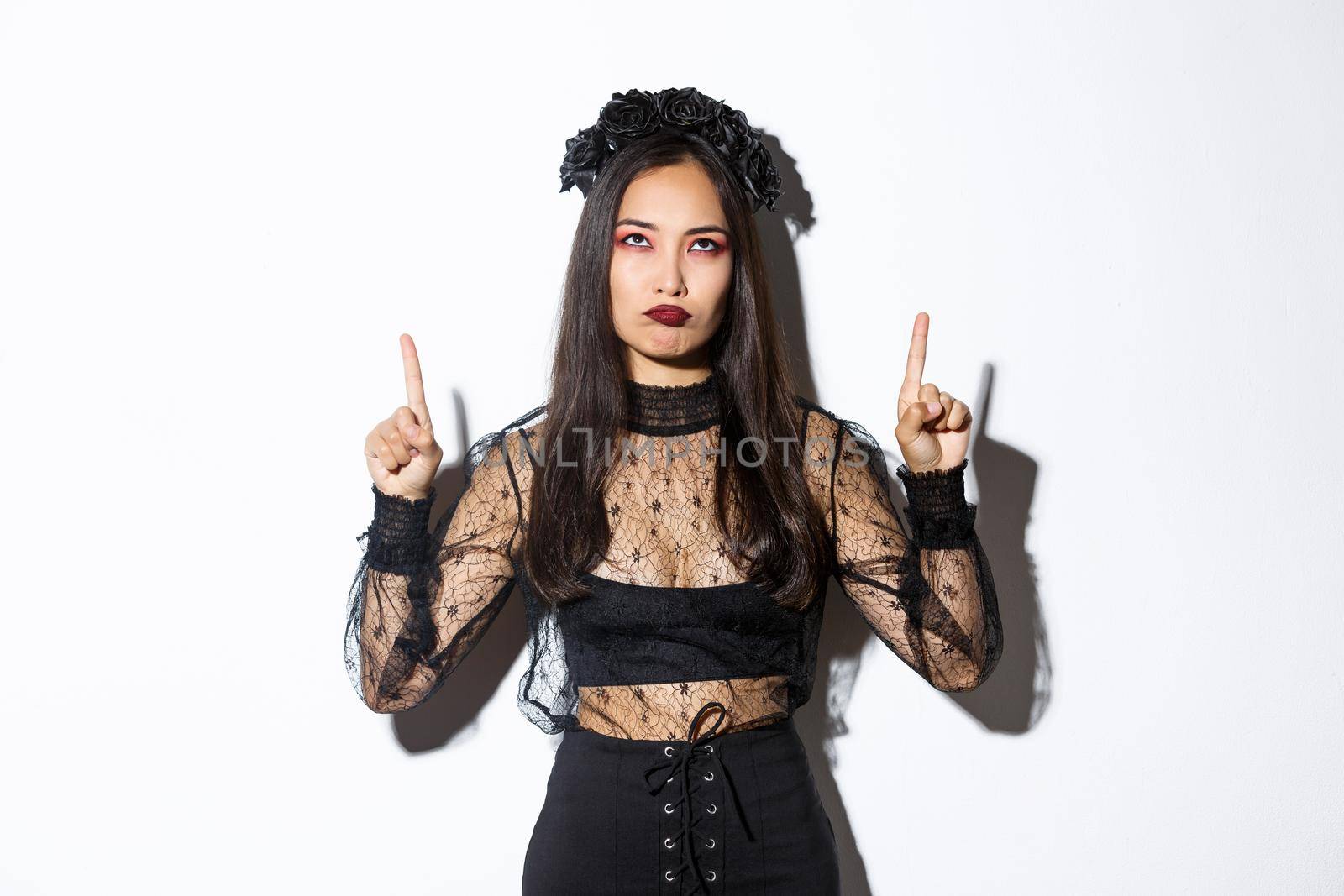 Grumpy asian female in witch costume looking disappointed and upset, pointing fingers up and sulking displeased, standing over white background in halloween costume by Benzoix