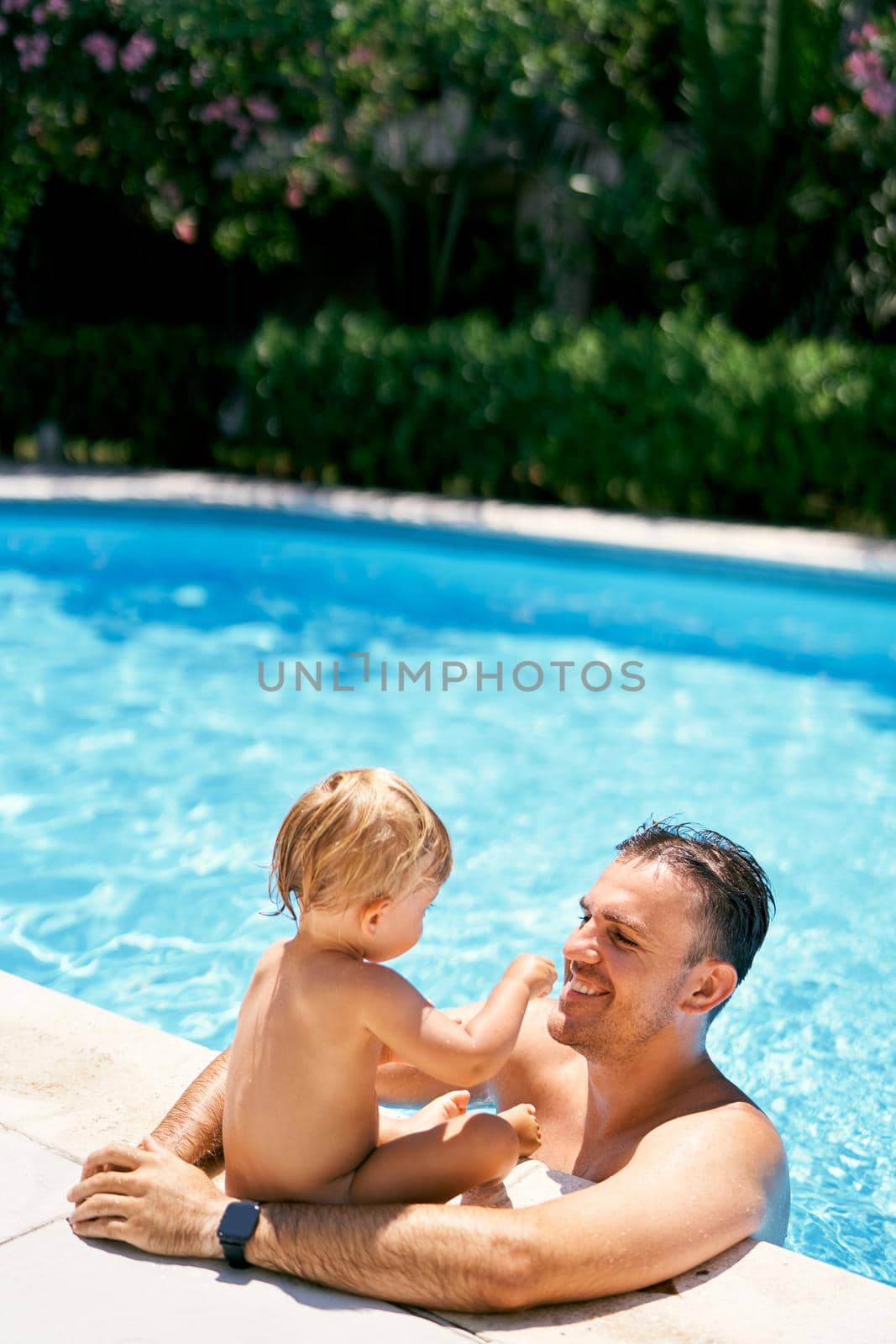 Smiling dad stands in the water and hugs a small child sitting on the edge of the pool by Nadtochiy
