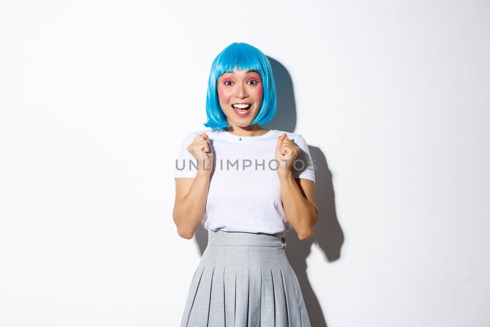 Portrait of excited beautiful asian girl looking hopeful, clenching fists and smiling amazed, standing over white background in blue wig.