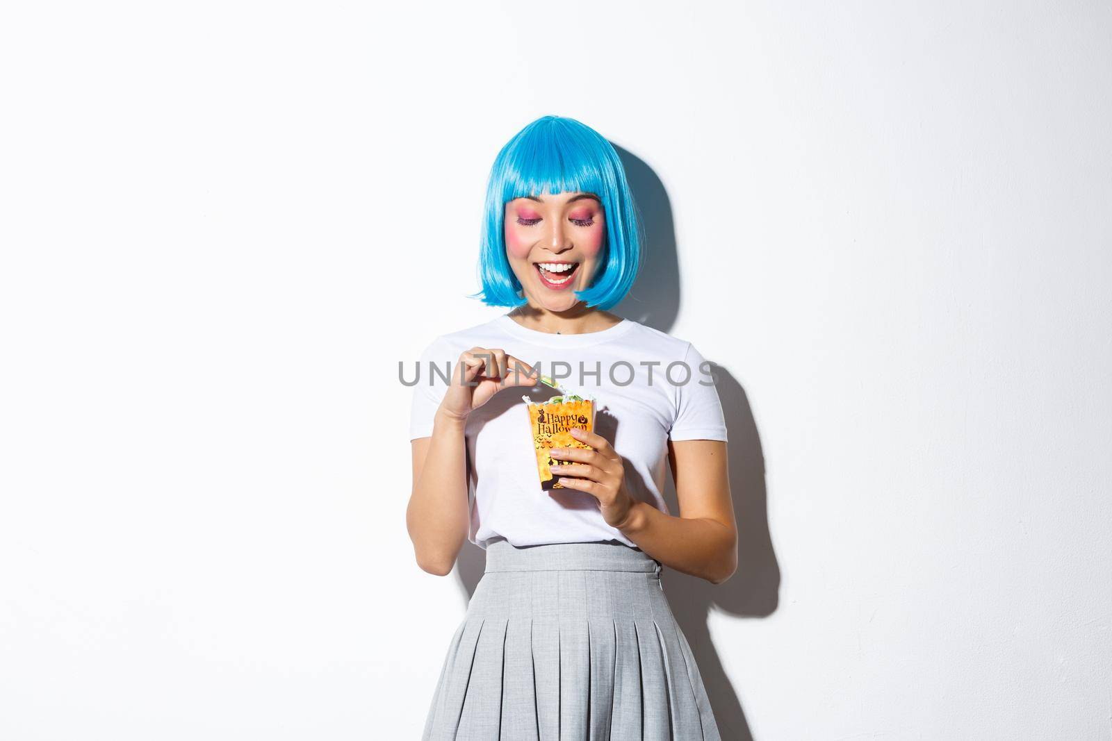 Image of cute asian girl celebrating halloween, eating sweets after trick or treat event, wearing blue short wig and anime schoolgirl costume, standing over white background.