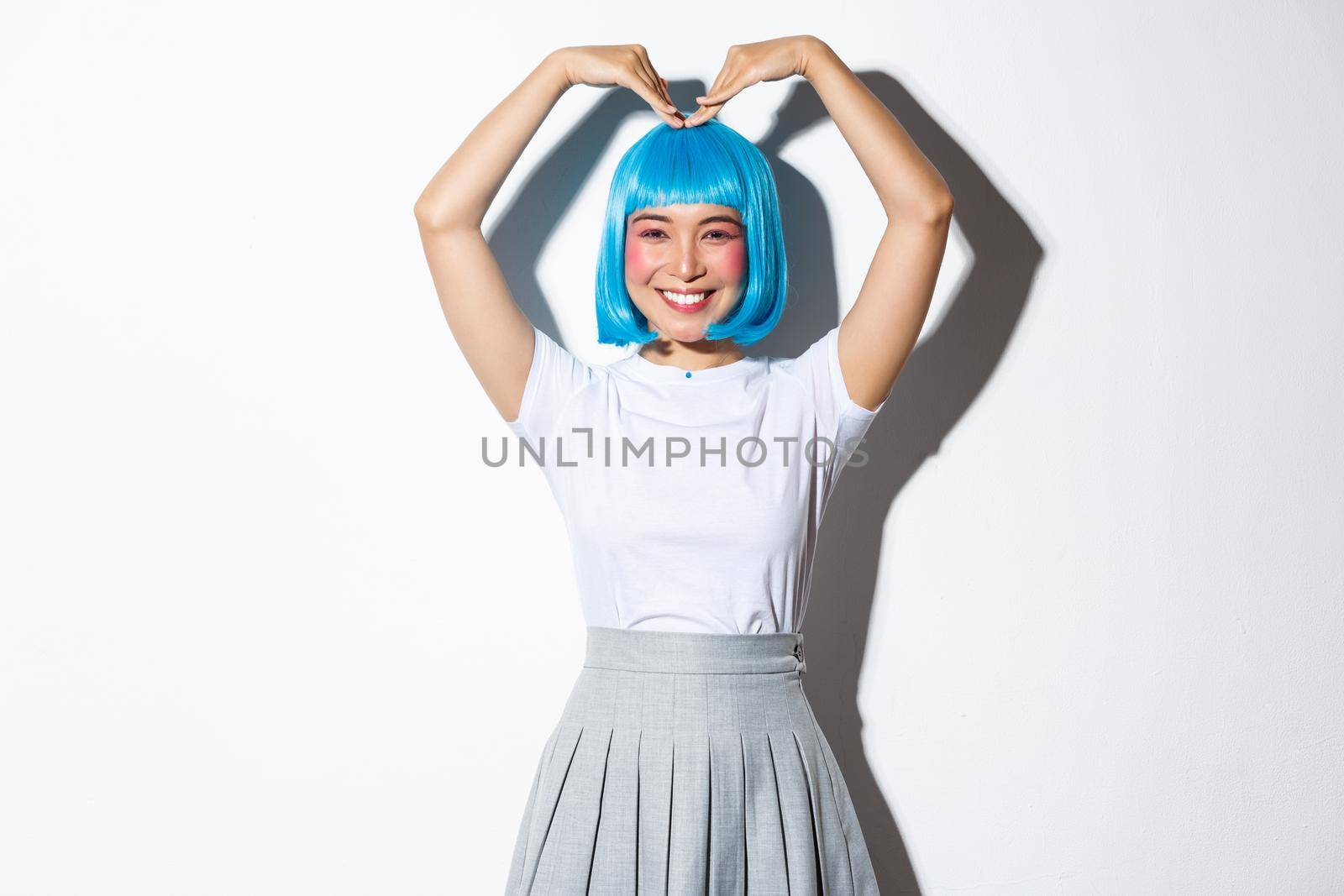 Portrait of cute japanese girl in blue wig showing heart gesture and smiling, standing over white background by Benzoix