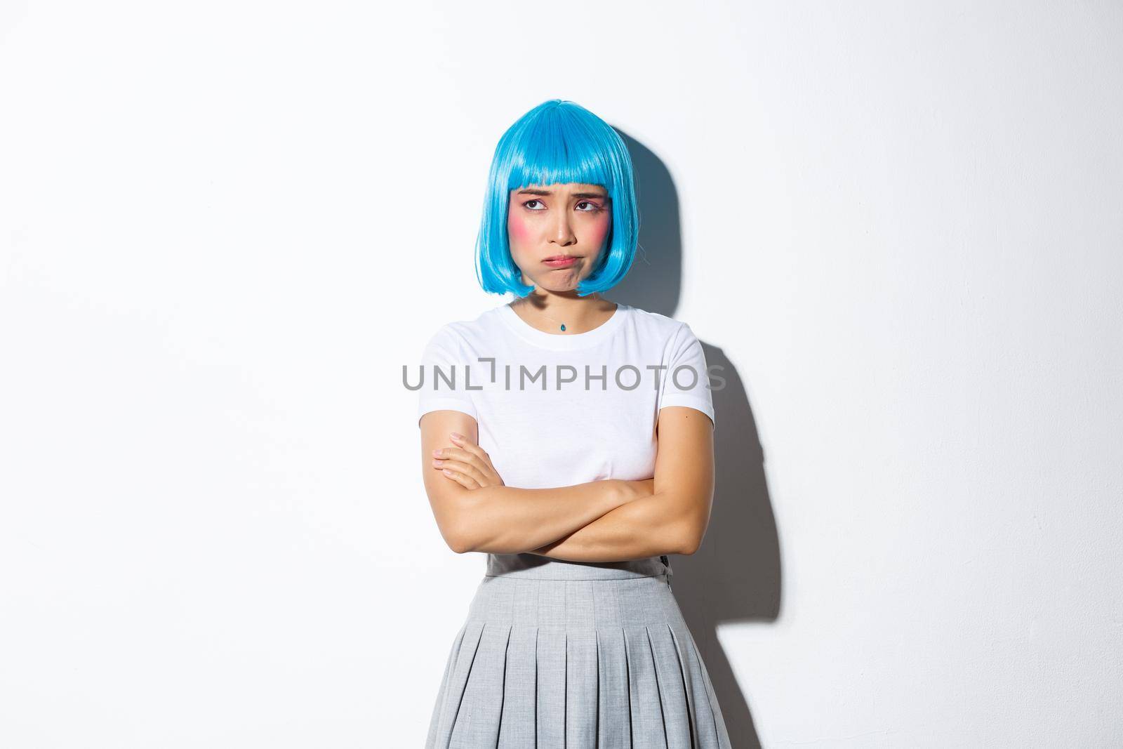 Portrait of cute asian girl looking troubled or upset, cross arms on chest and looking aside, standing in blue wig and school uniform, dressed for halloween celebration.