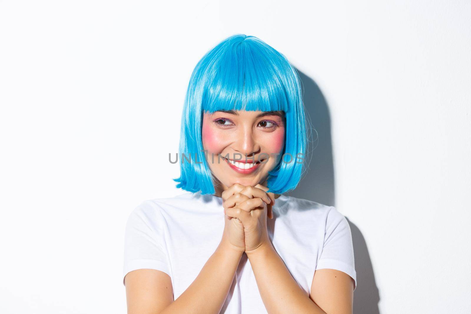 Close-up of adorable dreamy asian girl looking left and smiling, clasping hands together hopeful, wearing blue wig for halloween party, standing over white background.