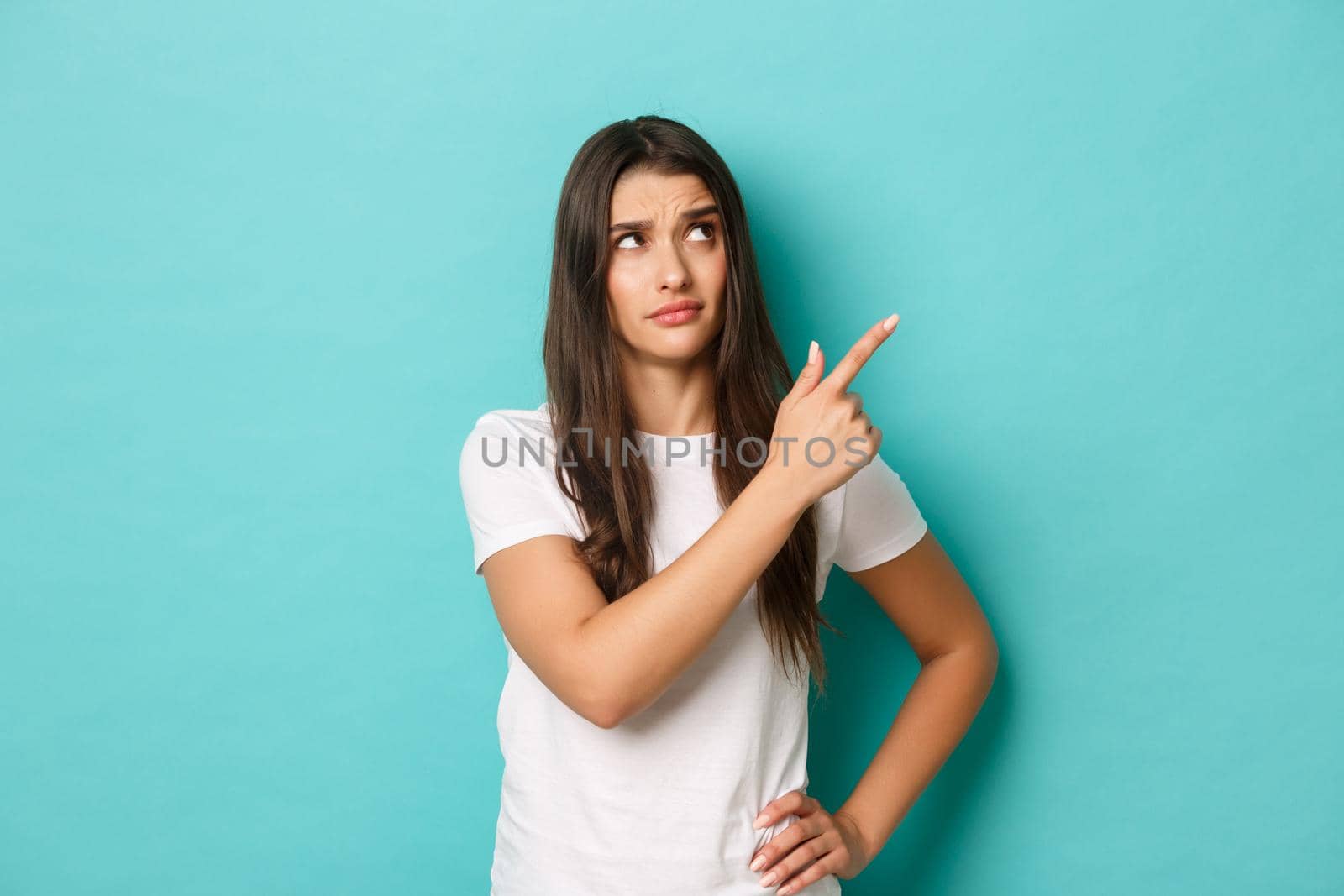 Image of doubtful brunette girl in white t-shirt, looking confused and something strange, pointing at upper right corner logo, standing over blue background.