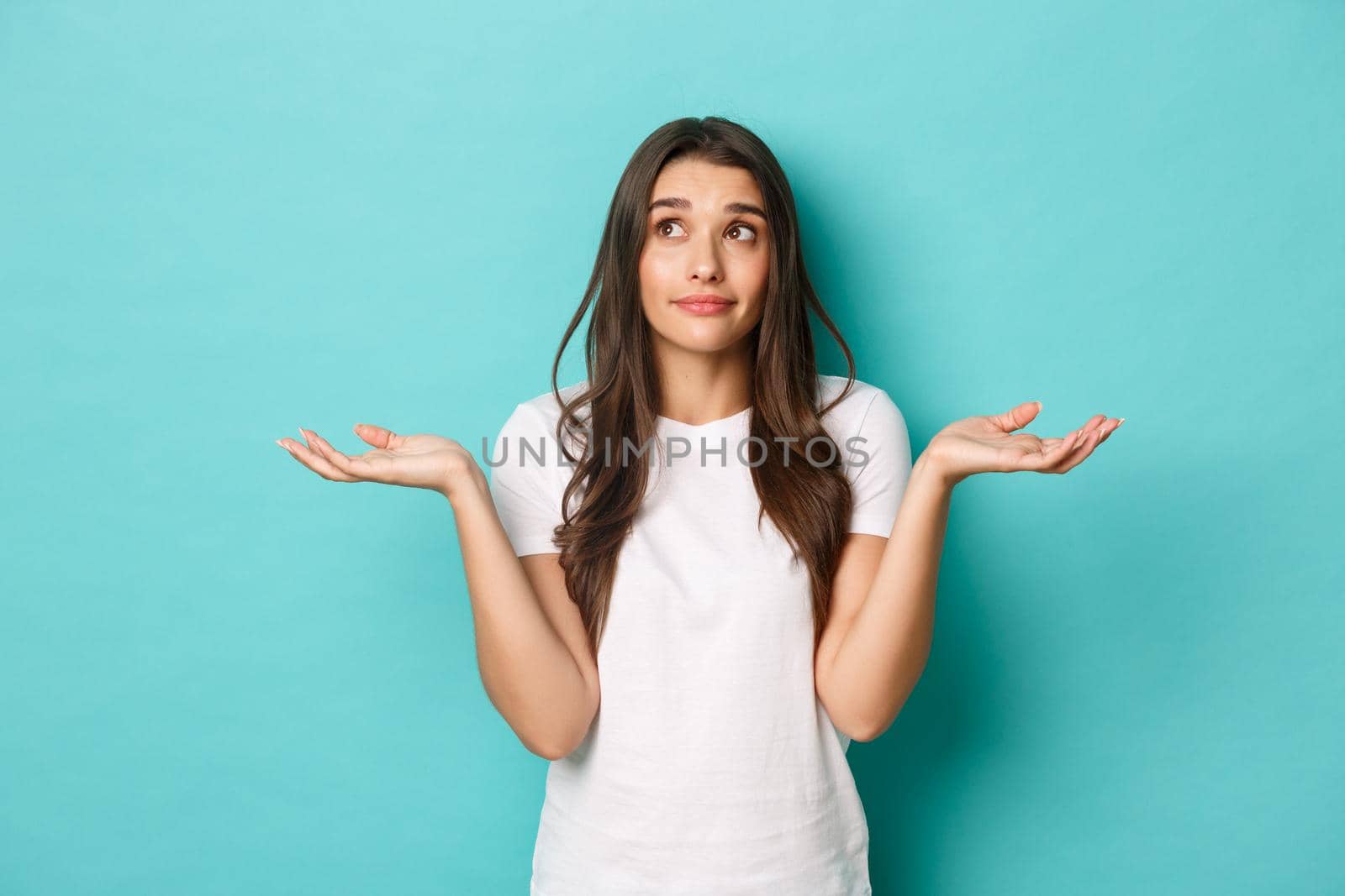 Image of pretty brunette woman in white t-shirt, dont know anything, shrugging and smiling clueless, standing over blue background.