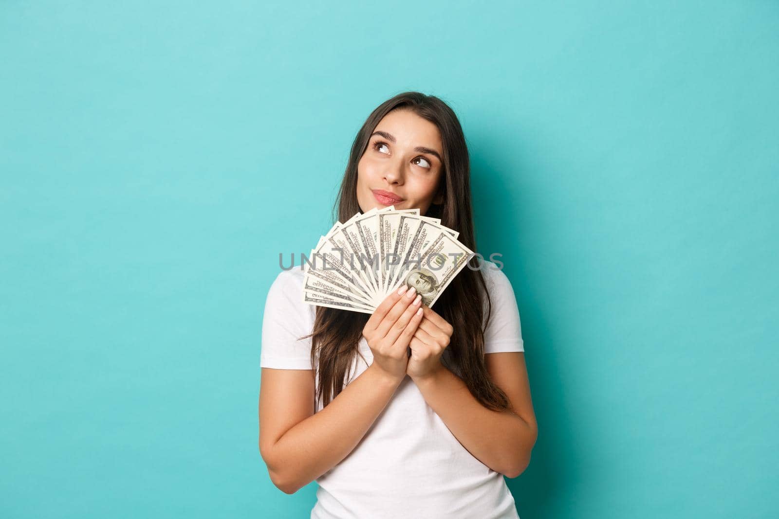 Portrait of attractive brunette woman in white t-shirt, dreaming about shopping, holding money, standing over blue background.