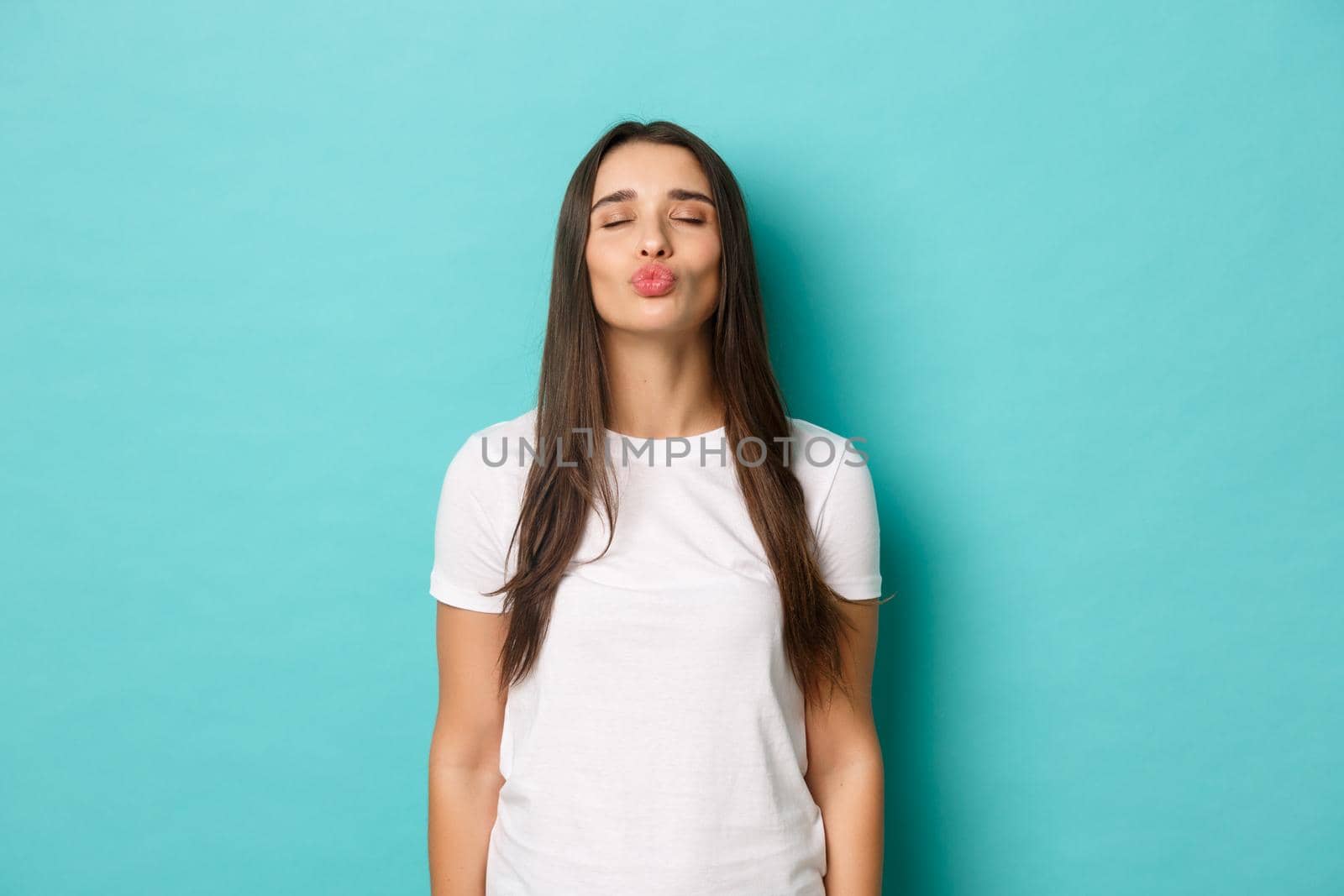 Portrait of cute girlfriend in white casual t-shirt, close eyes and pouting, waiting for kiss, standing over blue background.