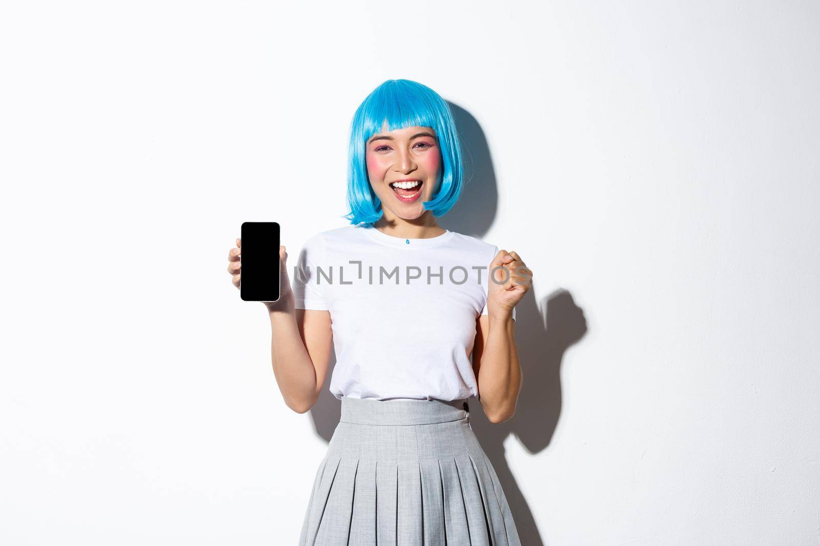 Image of cheerful winning asian girl, showing mobile phone screen and make fist pump gesture, smiling from rejoice, standing over white background.
