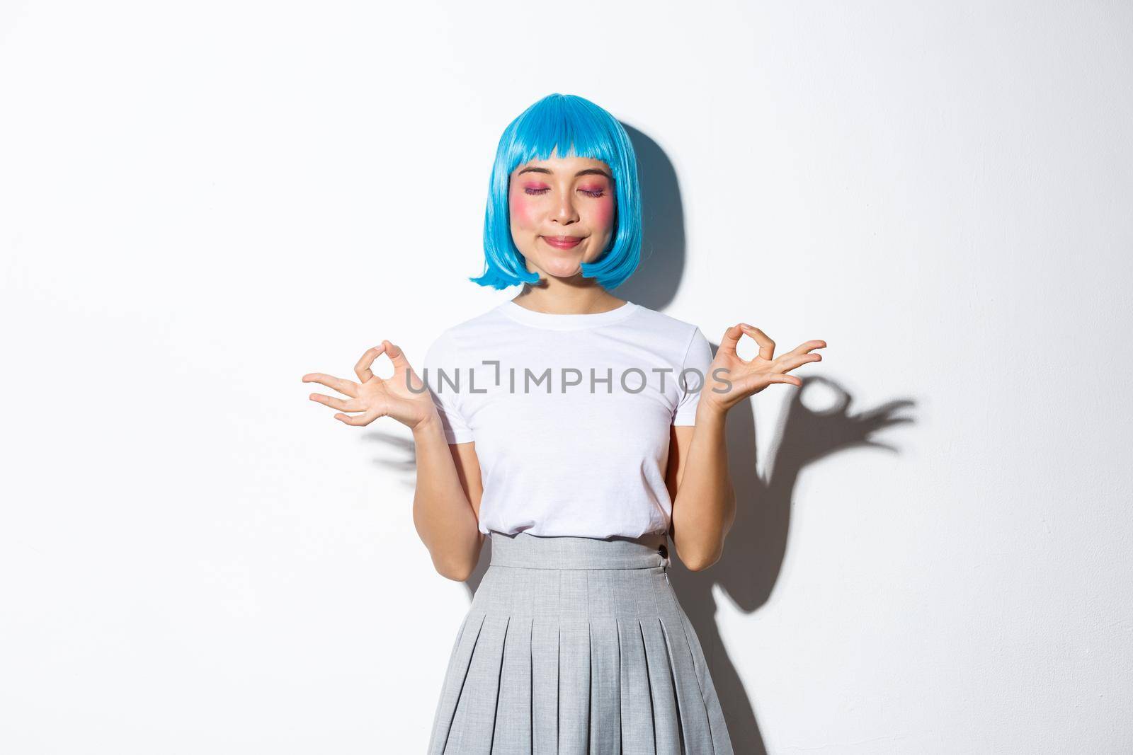 Portrait of beautiful asian girl in blue wig meditating, looking peaceful and smiling with eyes closed, standing in halloween costume over white background.