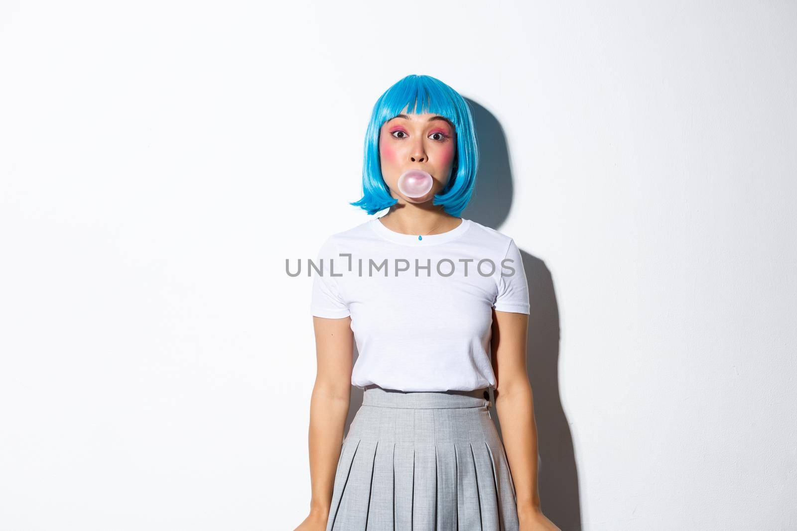 Beautiful asian girl in blue wig and schoolgirl costume, blowing bubble gym with silly face, dressed for halloween party as anime character, standing over white background.