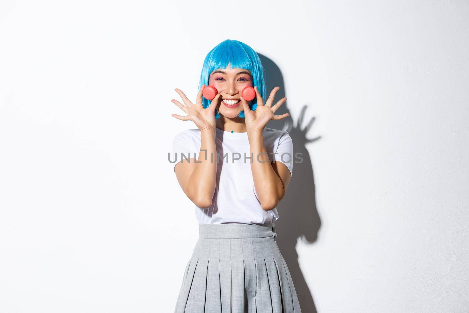 Image of beautiful smiling asian girl looking silly, showing macaroons, wearing blue anime wig, standing over white background.