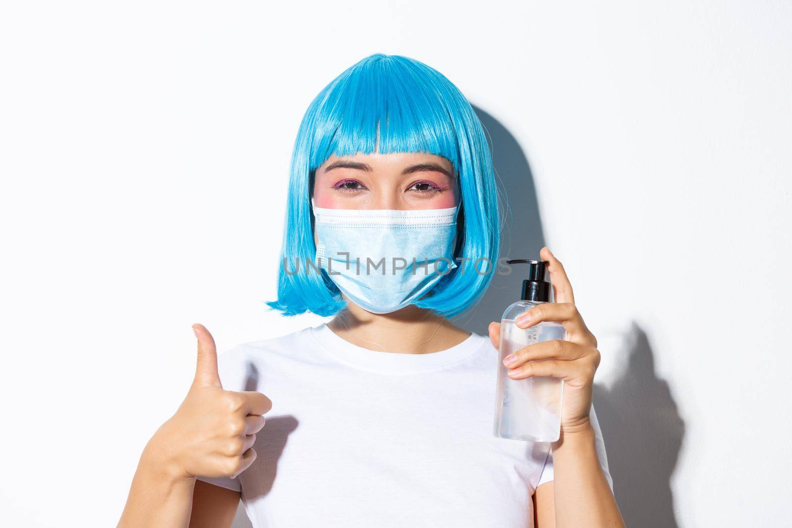 Concept of halloween celebration and coronavirus. Close-up of cute smiling asian girl in blue wig and medical mask showing hand saniziter and thumb-up.