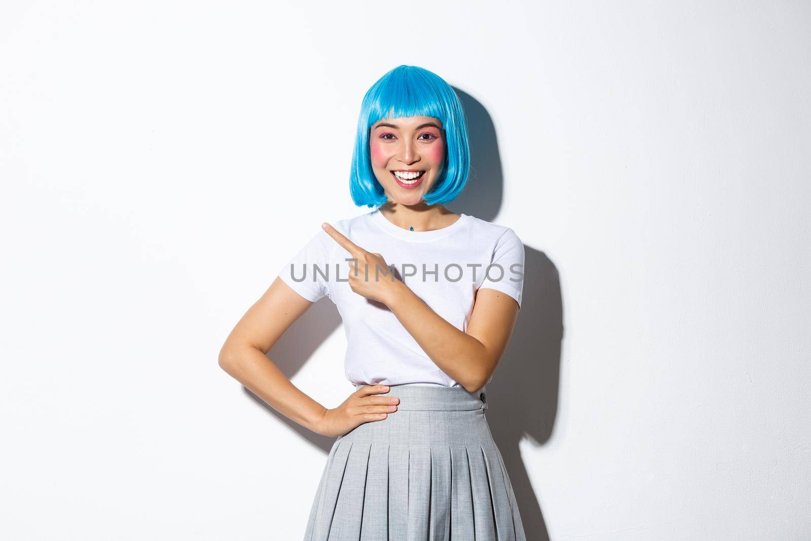 Cheerful asian girl smiling and pointing finger at upper left corner, showing your logo or banner over white blue background, wearing blue short wig for party or halloween.
