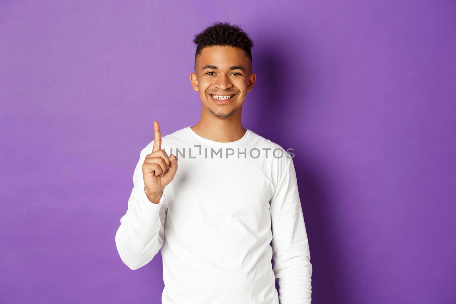 Image of cheerful african-american guy in white sweatshirt, showing one finger and smiling, standing over purple background.