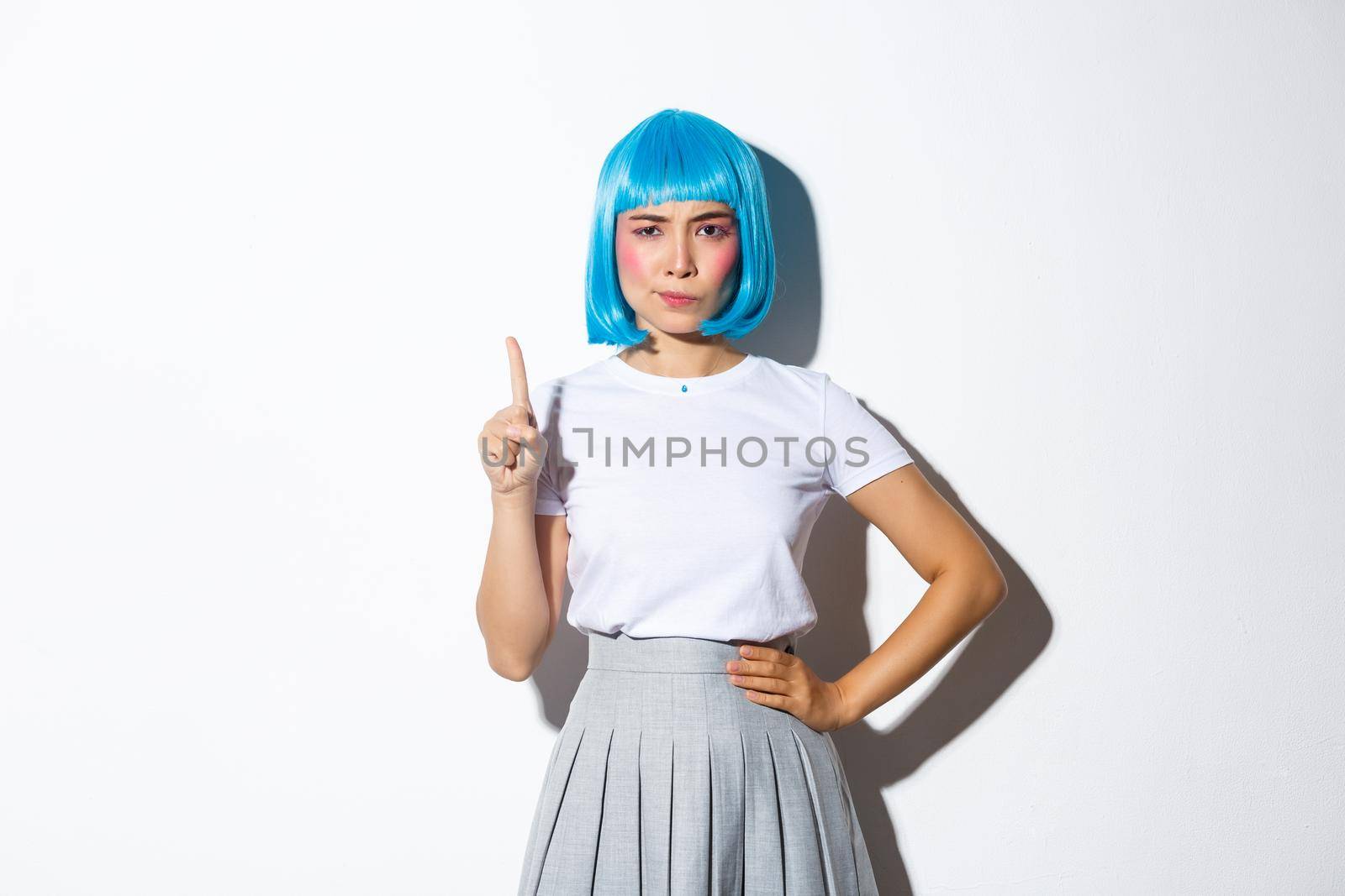 Portrait of serious asian girl in blue party wig looking disappointed, shaking finger and scolding someone bad behaviour, standing over white background.