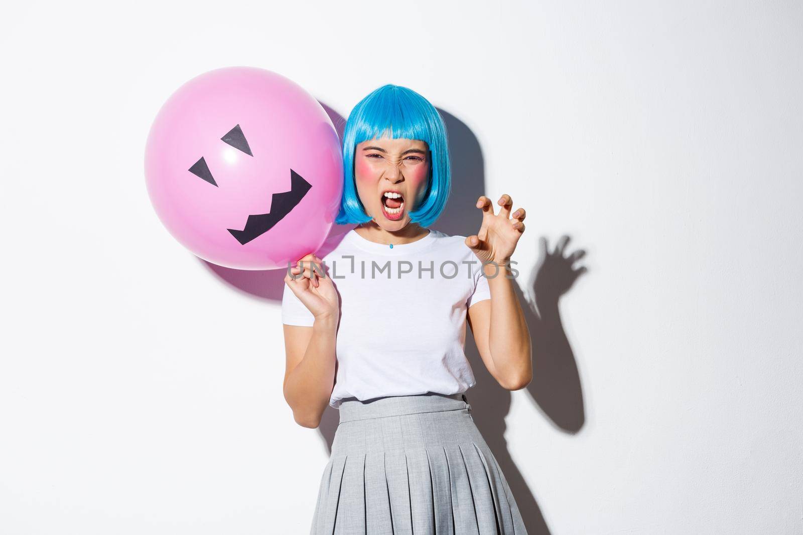 Portrait of funny asian girl trying to scare someone on halloween, wearing blue wig and holding pink balloon with scary face.