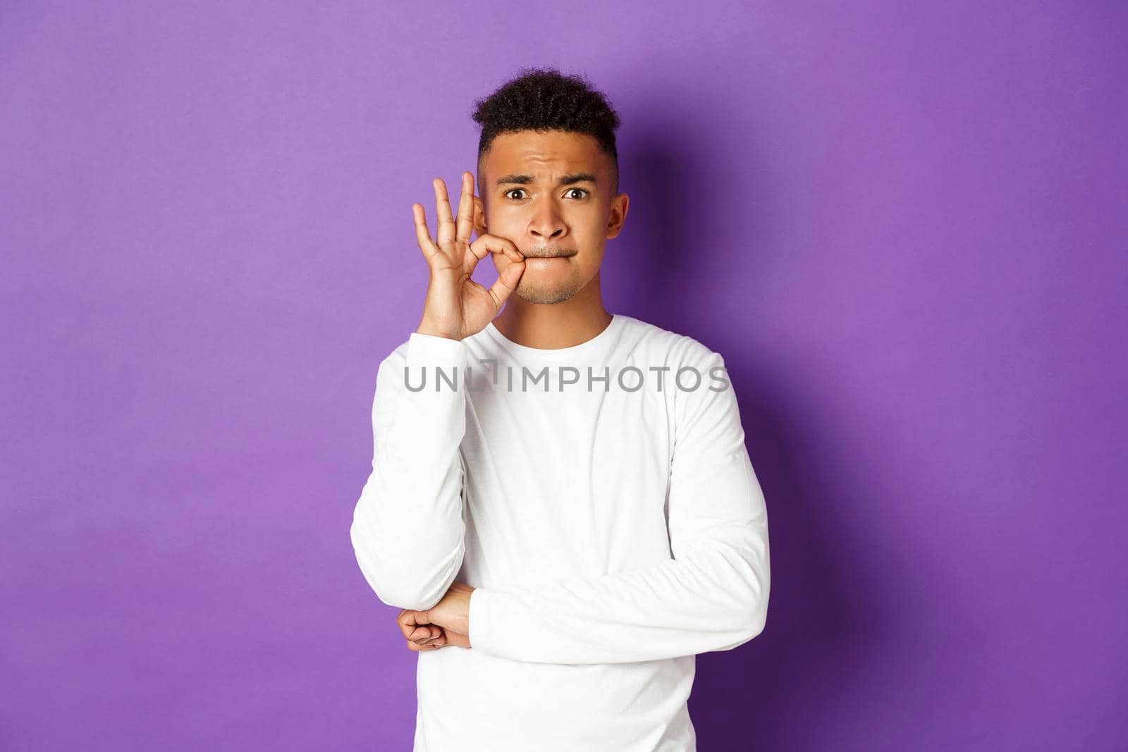 Image of african-american man in white sweatshirt, zipping mouth to make promise, seal lips, keeping secret, standing over purple background.