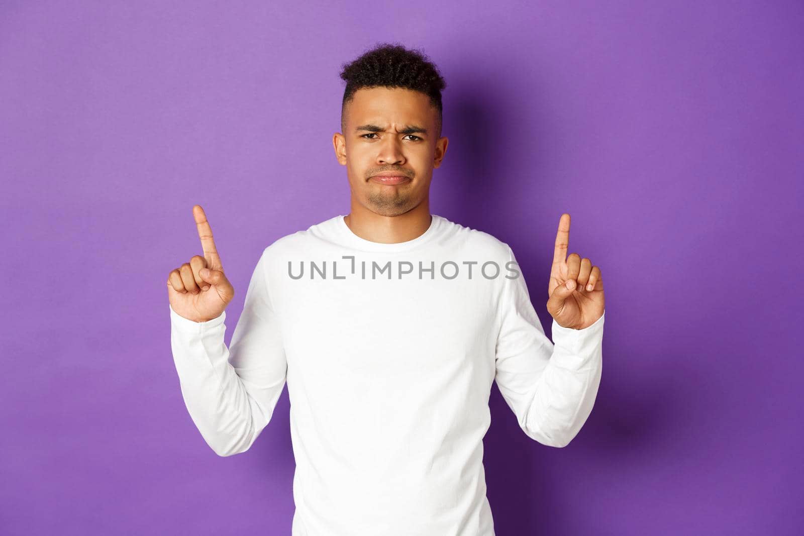 Displeased african-american guy in basic sweatshirt, grimacing and frowning disappointed, pointing fingers up at something bad, standing over purple background.