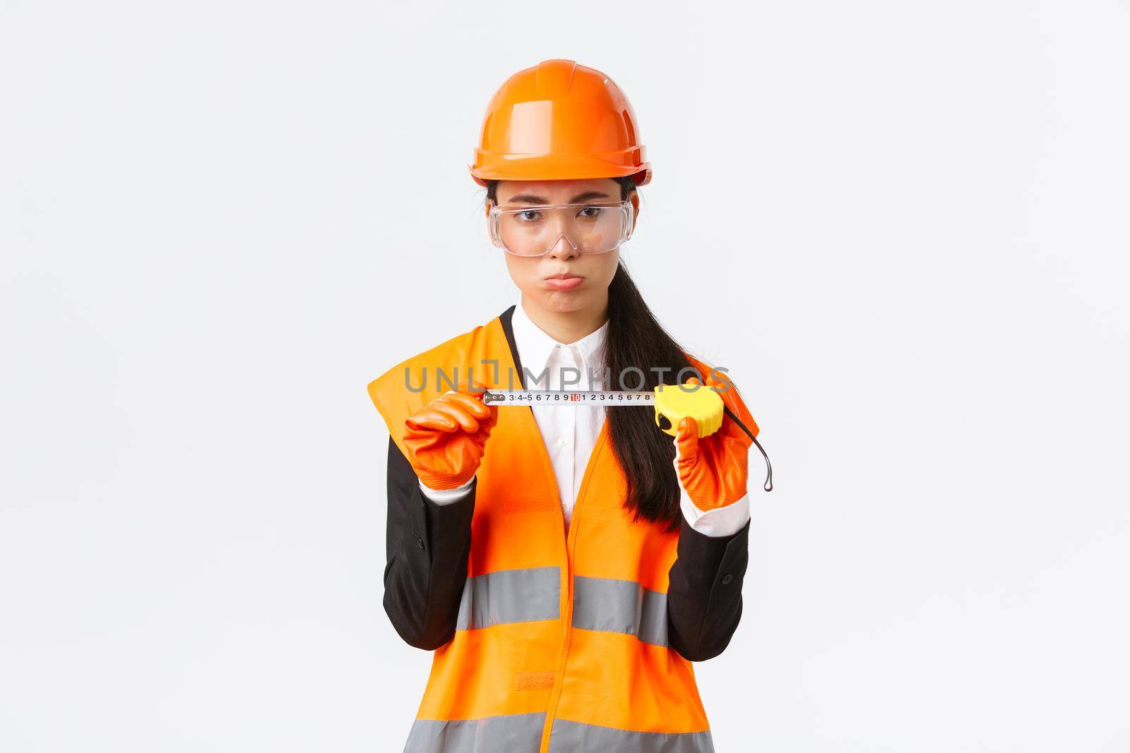 Disappointed gloomy female asian construction engineer, architect showing small size on tape measure, pouting upset with measurements, standing displeased in safety helmet, white background by Benzoix