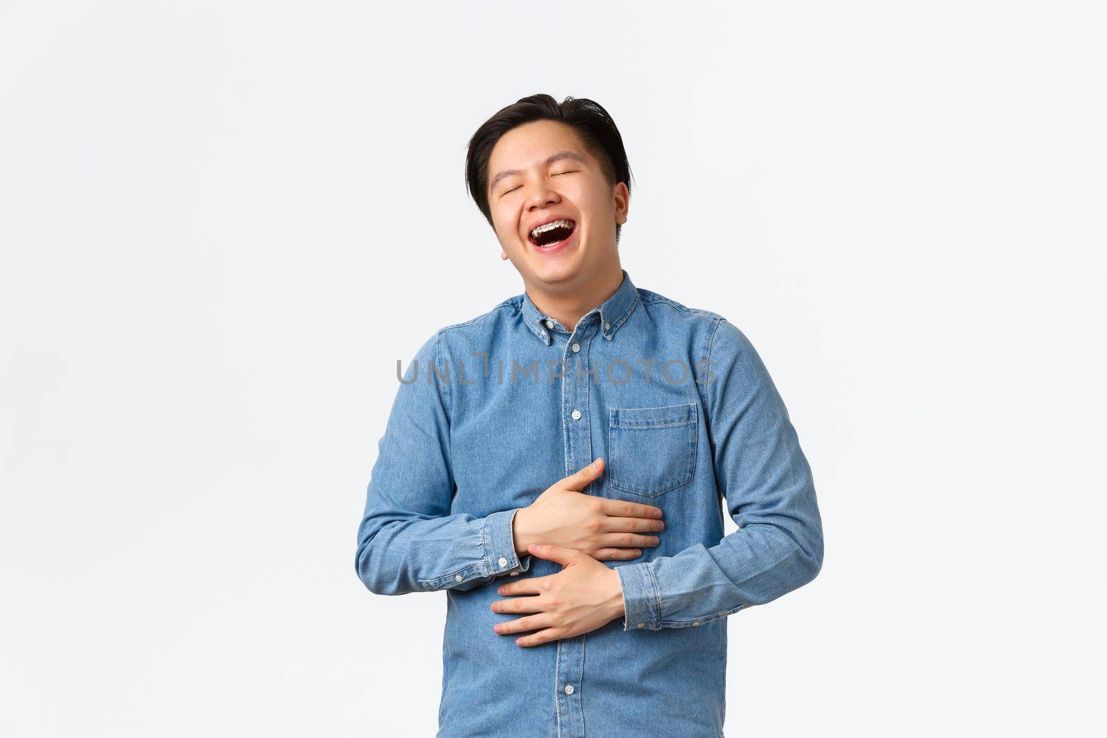 Carefree joyful handsome asian male student having fun, watching comedy, laughing out loud with closed eyes. Guy chuckle and touching belly, enjoying hilarious movie, white background.