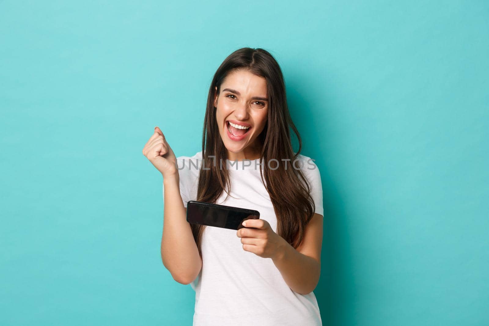 Image of happy young woman in white t-shirt, playing mobile phone game and winning, rejoicing while standing over blue background.