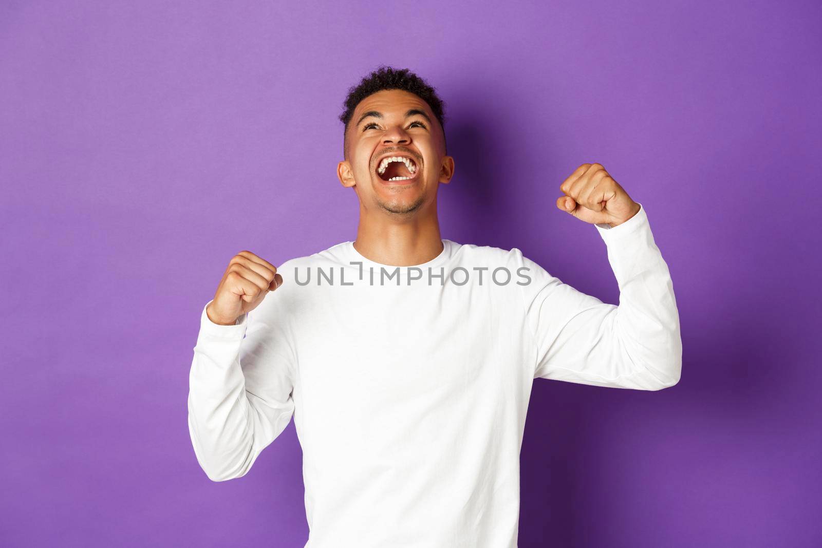 Image of happy and relieved african-american man, celebrating victory, looking up and making fist pump gesture, shouting yes with joyful expression, standing over purple background by Benzoix