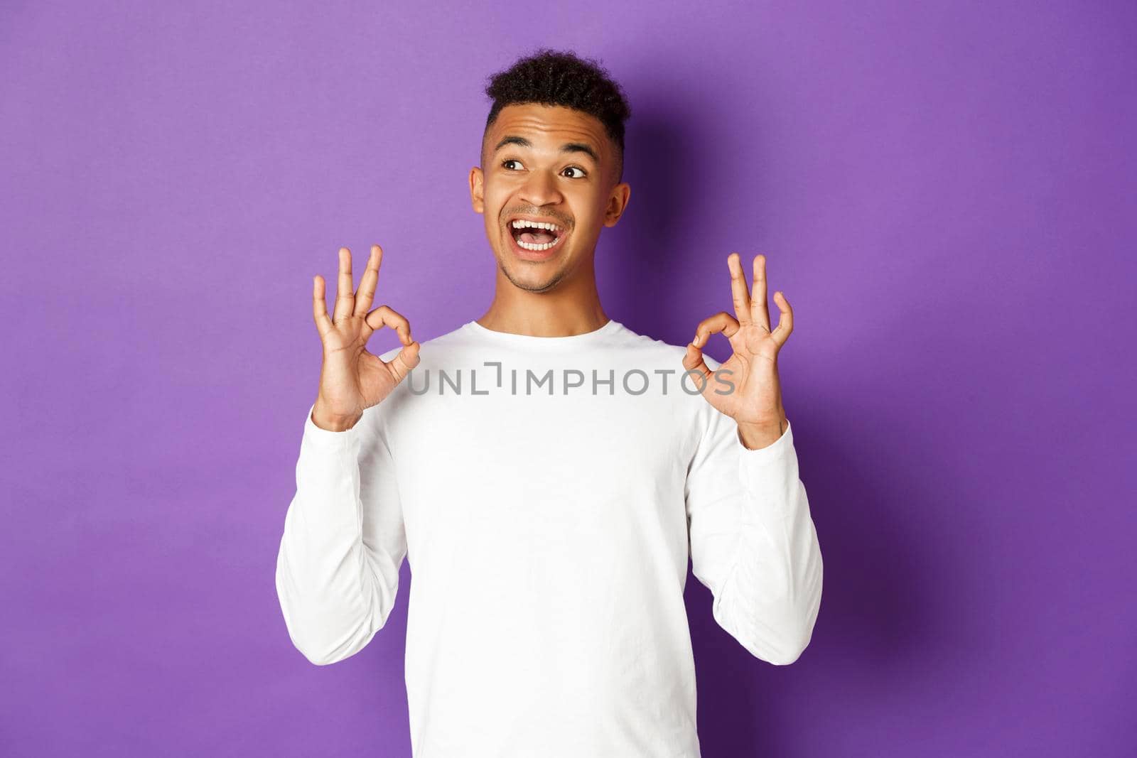 Image of satisfied african-american male student, looking pleased, looking at upper left corner and showing okay sign, standing over purple background.